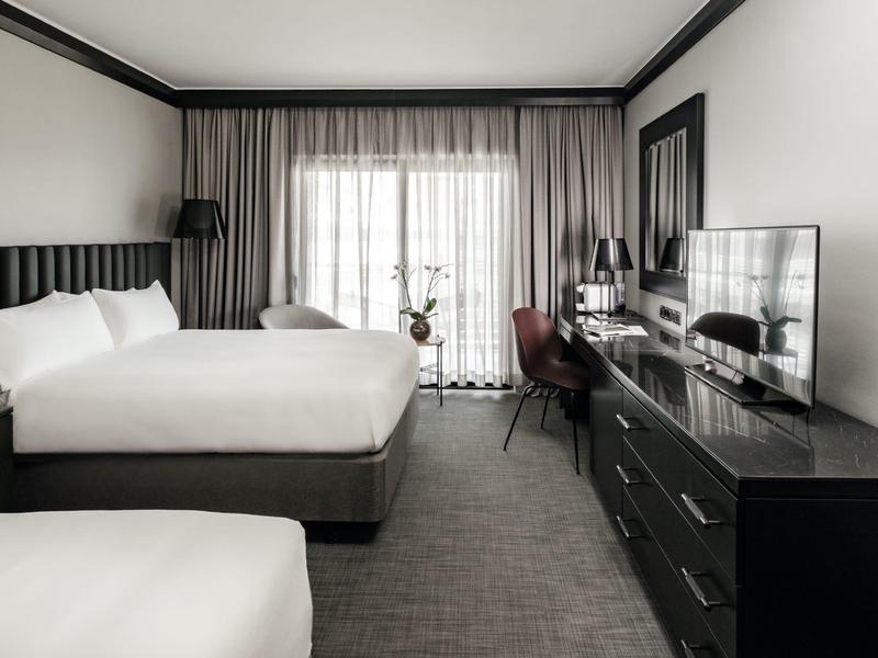 Modernes Hotelzimmer mit zwei großen Betten, Schreibtisch, Fernseher und großem Fenster mit Vorhängen.