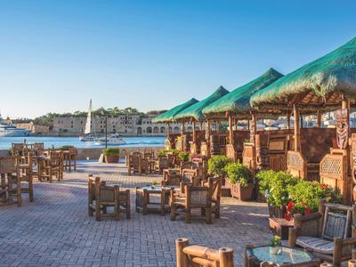 Restauracja na świeżym powietrzu z drewnianymi stołami i strzechą przy słonecznym porcie.