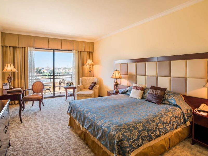 Jasny pokój hotelowy z dużym łóżkiem, wykładziną i balkonem z widokiem na miasto.