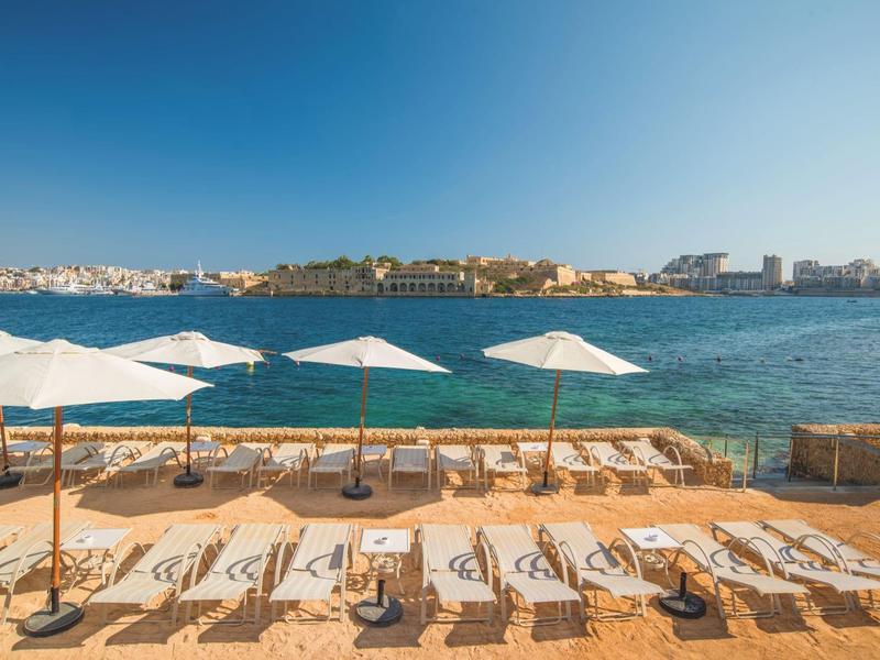 Słoneczny taras z leżakami i białymi parasolami nad wodą z widokiem na miasto.