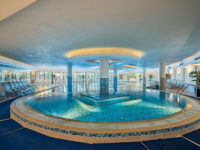 Jasny basen kryty o okrągłym kształcie, ze szklanymi ścianami, otoczony leżakami i roślinami.