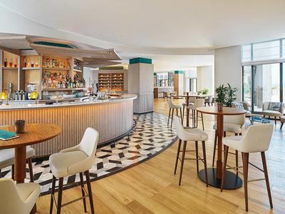 Lichte hotelbar met houten en mozaïekvloer, hoge tafels en gestoffeerde stoelen.