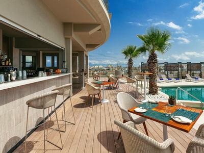 Terras met bar, tafels en uitzicht op zwembad en stad onder blauwe lucht.