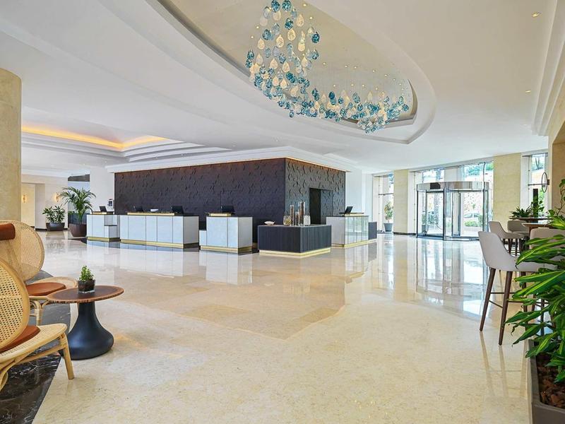 Großer heller Hotel-Eingangsbereich mit Rezeption, Sitzgelegenheiten und Pflanzen an der Seite.