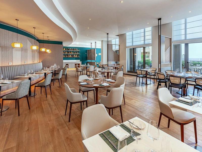 Moderner Restaurantraum mit Holzboden, bequemen Stühlen, großen Fenstern und hellen Wänden.