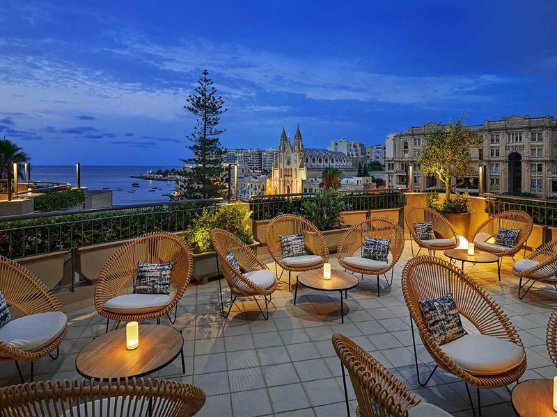 Dachterrasse mit gemütlichen Korbsesseln, beleuchteten Tischen und Blick auf historische Gebäude bei Abenddämmerung.