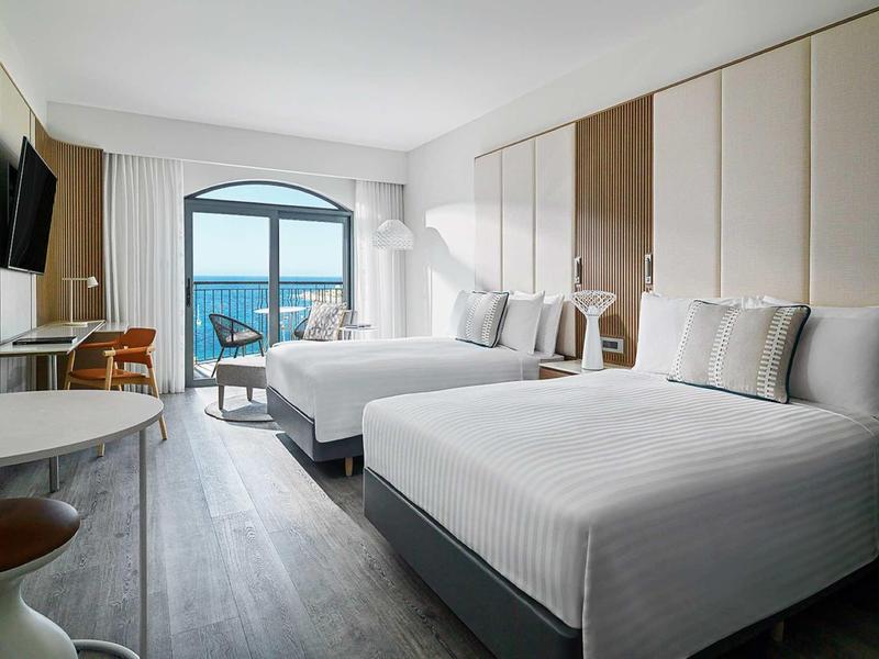 Helles Hotelzimmer mit zwei großen Betten, Balkon mit Meerblick und modernem Interieur.