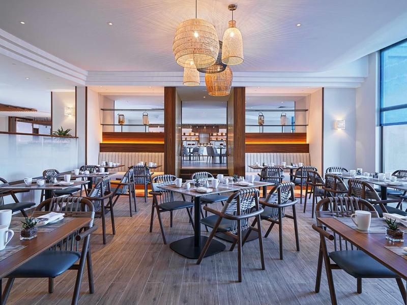 Modernes Restaurant mit Holzstühlen, runden Tischen, großen Fenstern und warmen Deckenlampen.