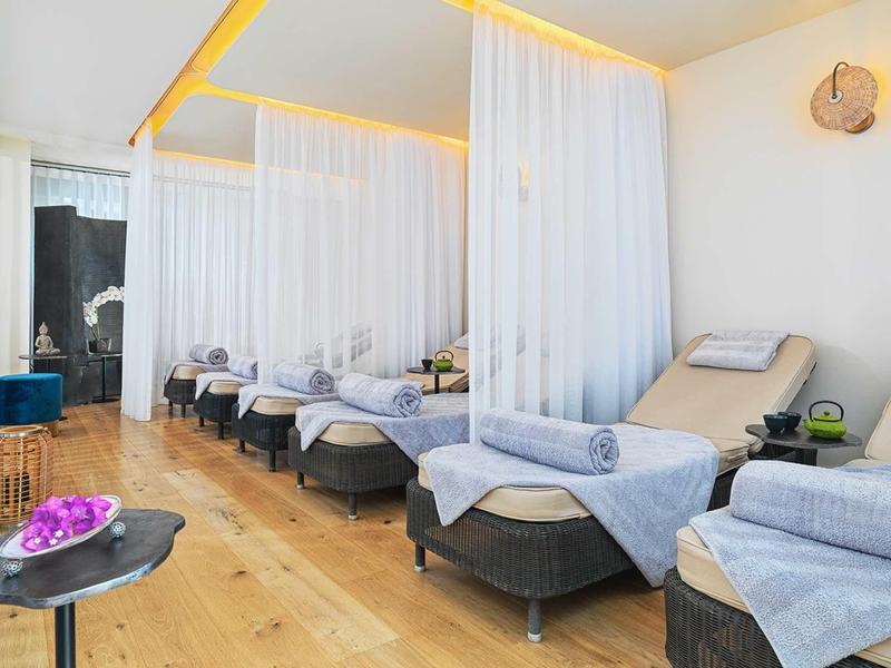 Heller Wellnessraum mit Liegen, weißen Vorhängen, Holzboden und entspannender Atmosphäre.