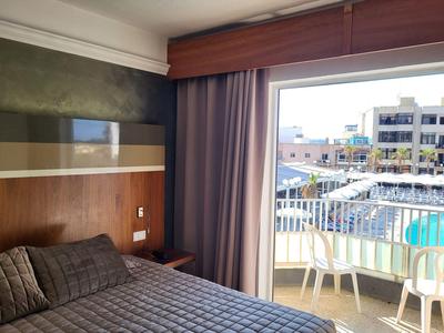 Hotelzimmer mit Doppelbett, Holzpaneelen und Balkon mit Blick auf Pool und Gebäude.