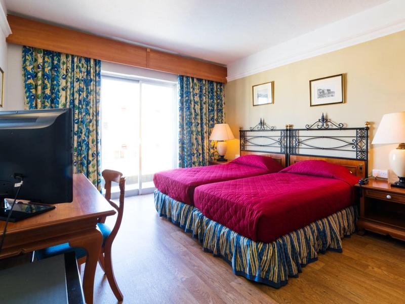 Světý hotelový pokoj se dvěma jednolůžky, televizí, stolem a dveřmi na balkon.