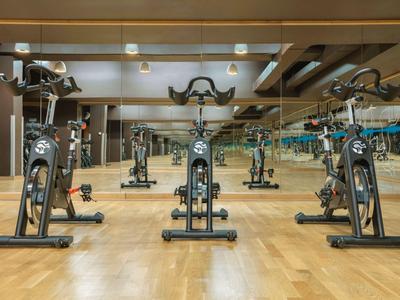 Tre cyclette in una palestra con pavimento in legno e grandi specchi a parete.