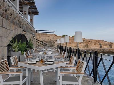 Tavoli da pranzo su una terrazza con vista sul mare e scogli rocciosi sullo sfondo.