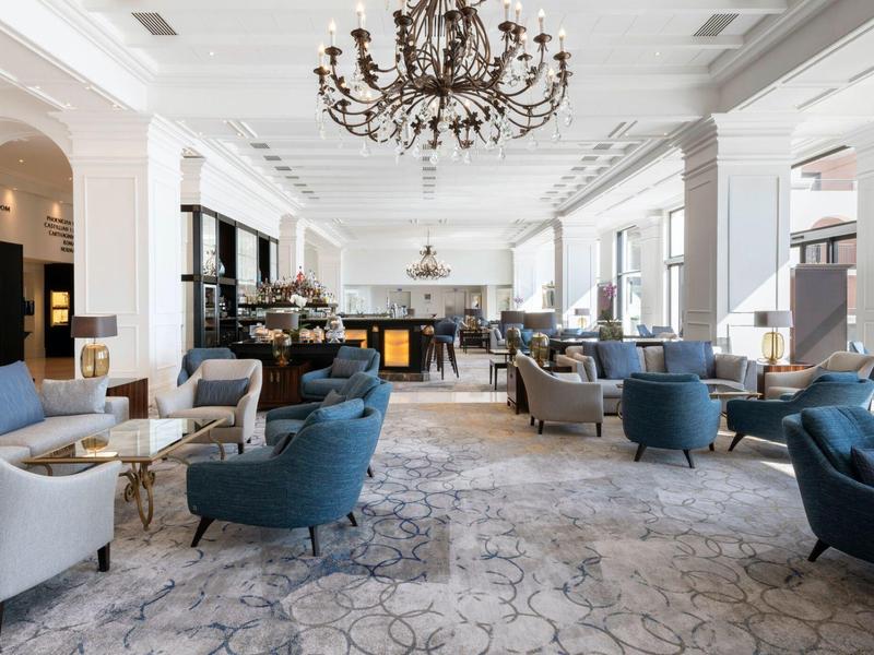 Helles, elegantes Hotel-Lounge mit blauen und grauen Sesseln, Kronleuchter und großen Fenstern.