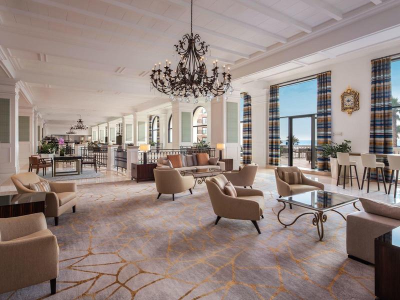 Helles, modernes Hotel-Lounge mit bequemen Sesseln, Kronleuchtern und großen Fenstern mit Blick auf Palmen.