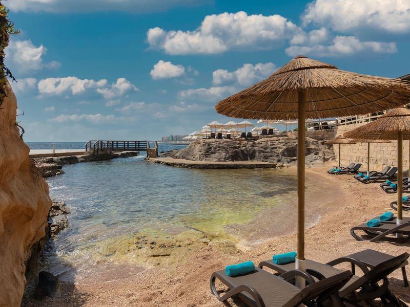 Sonnenschirme und Liegen am felsigen Strand mit klarem Wasser und blauem Himmel.