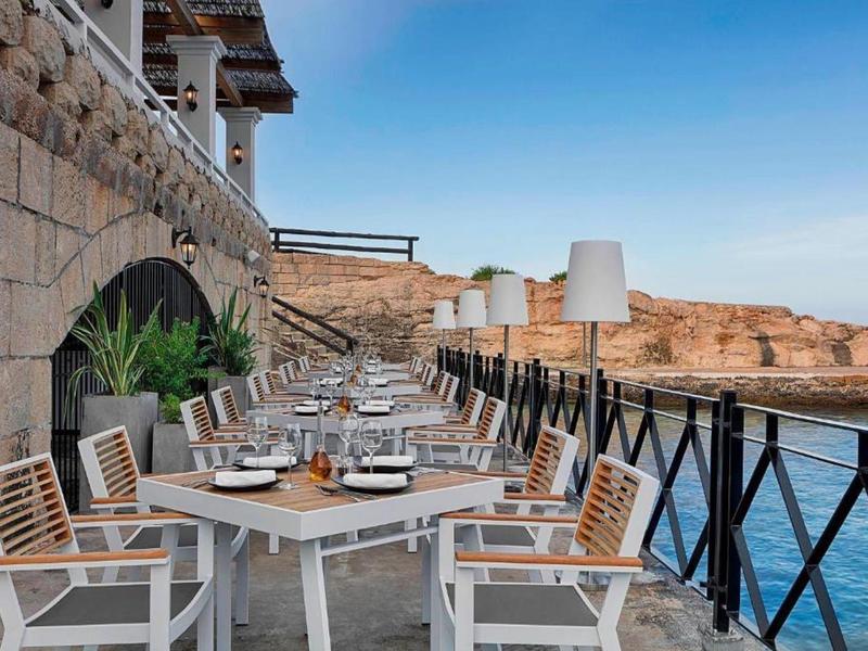Terrasse eines Restaurants mit Tischen und Stühlen am Meer mit Blick auf Felsen unter klarem Himmel