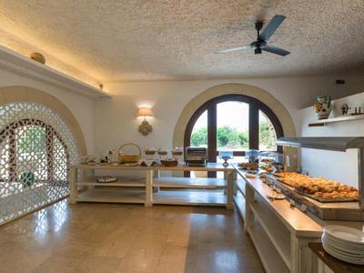 Sala de desayuno luminosa con buffet, mesas de madera y grandes ventanas arqueadas con vista a la vegetación.