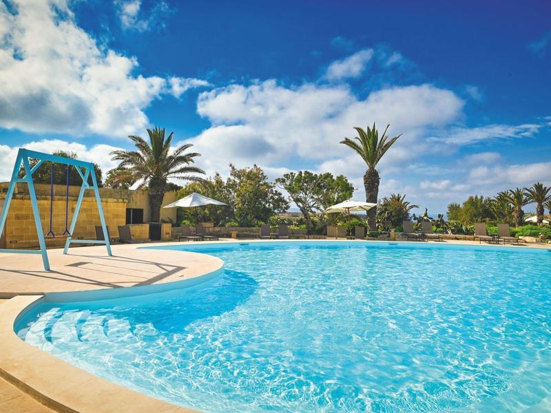Gran piscina al aire libre con agua azul rodeada de palmeras bajo un cielo azul.