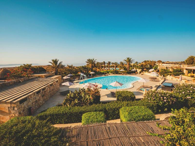 Piscina redonda del hotel rodeada de tumbonas, palmeras y edificios bajo un cielo azul.