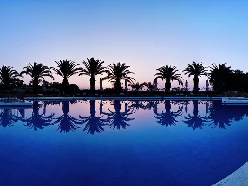 Piscina con palmeras reflejadas al atardecer en un resort hotelero.