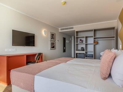 Moderní hotelový pokoj s manželskou postelí, televizí na zdi, psacím stolem a otevřenou skříní.