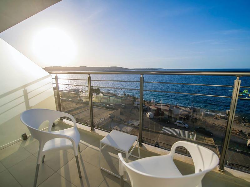 Balkon se dvěma bílými židlemi a malým stolkem s výhledem na moře za západu slunce.