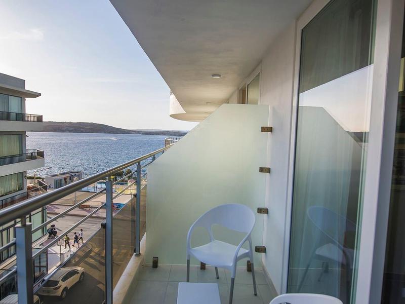 Balkon s bílou židlí, skleněným zábradlím a výhledem na moře za jasné oblohy.