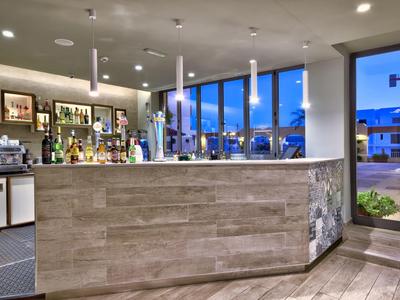 Área moderna de bar de hotel con mostrador de madera, estantes con bebidas y grandes ventanales al anochecer.