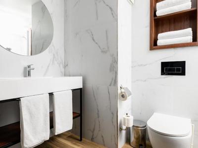 Baño de hotel moderno con inodoro suspendido, lavabo blanco y estante de madera con toallas.