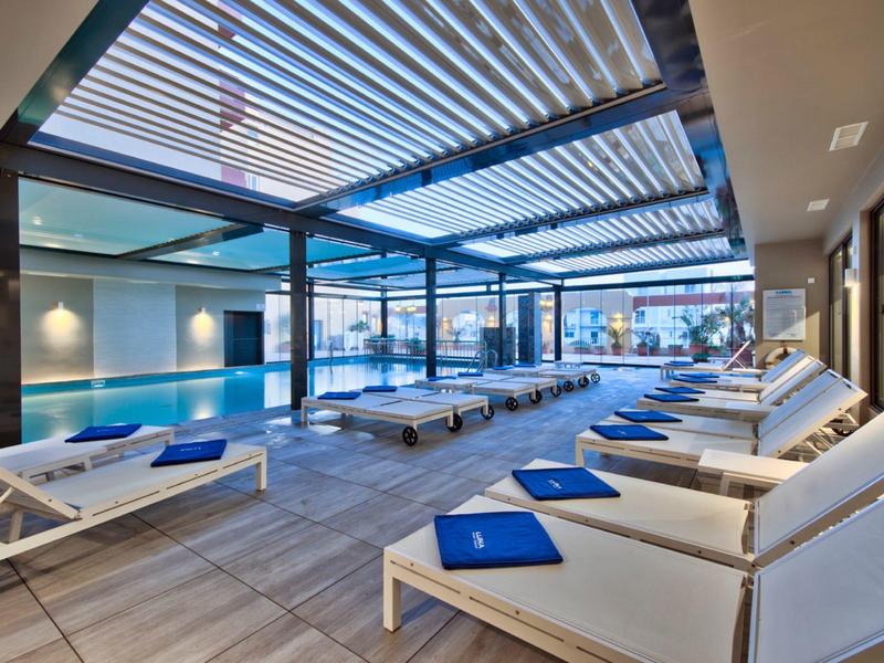 Piscina cubierta moderna con cómodas tumbonas y paredes de cristal que dan a una terraza con vistas a la ciudad.