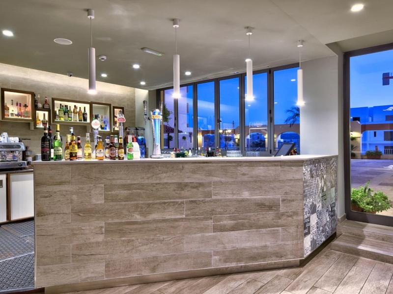 Área moderna de bar de hotel con mostrador de madera, estantes con bebidas y grandes ventanales al anochecer.