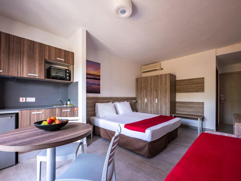 Habitación de hotel moderna con dos camas, kitchenet y mesa de comedor con frutero.