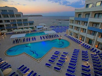 Blick auf einen Hotelpool mit Liegestühlen und Meer im Hintergrund bei Sonnenuntergang.