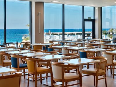 Heller Speisesaal mit vielen Tischen und Stühlen, Blick auf das Meer durch große Fenster.