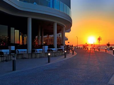 Abendliche Strandpromenade mit modernem Gebäude und Sonnenuntergang im Hintergrund.