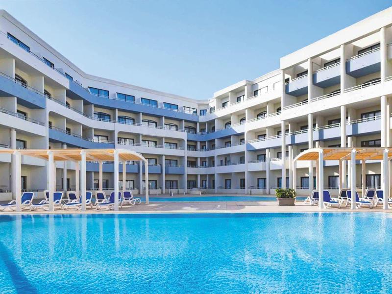 Modern hotelcomplex met groot blauw zwembad en parasols bij heldere lucht.