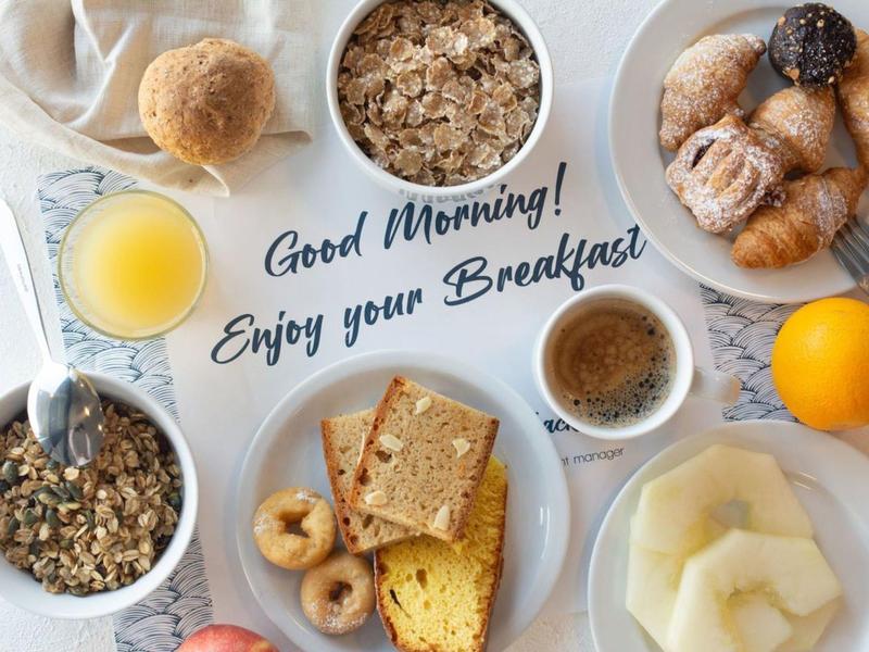 Ontbijttafel met sap, ontbijtgranen, koffie, gebak en fruit, met tekst 'Good Morning! Enjoy your Breakfast'.