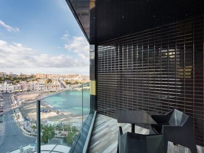 Balcony with glass railing, chairs, and table overlooking harbor and city.