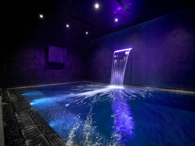Dark wellness room with illuminated indoor pool and waterfall wall.