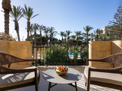 Balkon s proutěnými křesly s výhledem na bazén obklopený palmami pod jasnou oblohou.