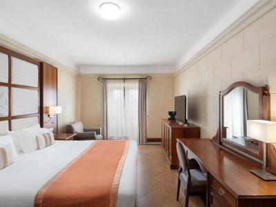 Světle hotelový pokoj s velkou postelí, psacím stolem, televizí a závěsy u okna.