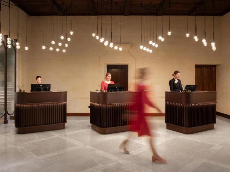 Hotelová recepce se třemi přepážkami a zaměstnanci, žena v červených šatech prochází kolem.