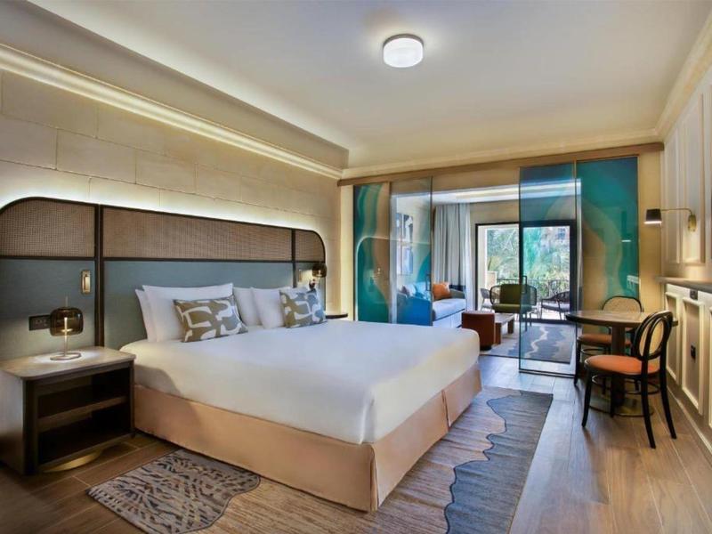 Moderní hotelový pokoj s velkou postelí, židlemi, koberci a balkónem s výhledem do zeleně.