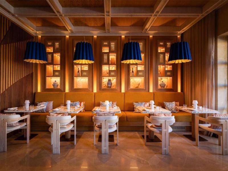 Útulná restaurace s dřevěným nábytkem, teplým osvětlením a dekorativními nástěnnými policemi.