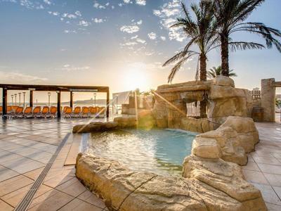 Poolbereich mit Felsenwasserfall und Sonnenuntergang hinter Palmen auf der Terrasse.