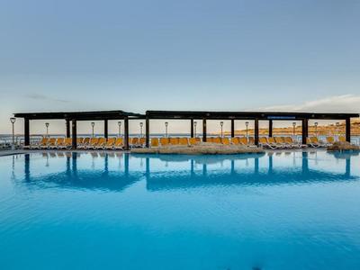 Velký venkovní bazén s moderními stínícími konstrukcemi a žlutými lehátky pod jasnou oblohou.