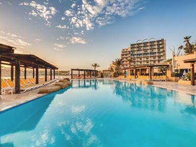 Grande piscina all'aperto con ombrelloni e edificio dell'hotel sullo sfondo al tramonto.