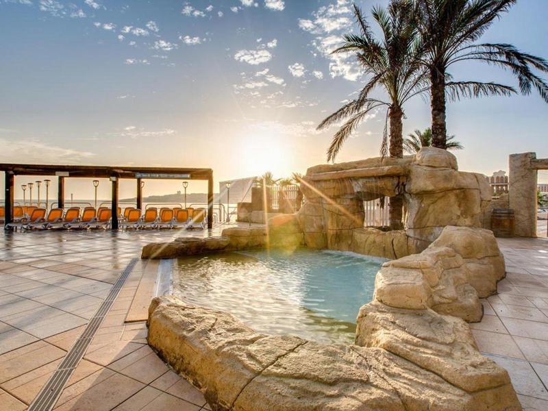 Bazénová oblast s skalním vodopádem a západem slunce za palmami na terase.