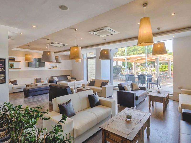 Světlý hotelový salonek s pohodlnými pohovkami, dřevěnými stolky a velkými okny směrem na venkovní prostor.